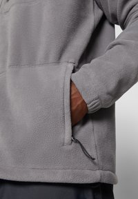 Hall grey fliisist pullover, millel on tekstureeritud pind, ees on tasku ja hemmi juures reguleeritav nöör. Pehme ja mugav materjal.