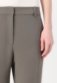 Grå bukser med en glat tekstur, der har en lige snit, for-pleats og en bæltestrop. Hånd hviler på siden.