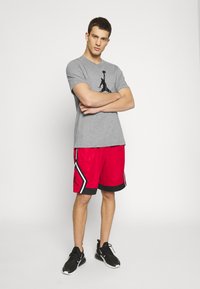 Camiseta gris con un logo negro, pantalones cortos rojos con acentos en negro y blanco, y zapatos deportivos negros. Postura concentrada, fondo neutral.
