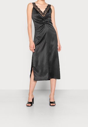 Vrouw draagt een zwarte satijnen midi-jurk met kanten rand, gerimpeld aan de voorkant, zijsplit en zwarte open-toe hoge hak sandalen, staand op een grijze vloer.