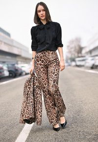 Pantaloni a stampa leopardata in beige e marrone, abbinati a una blusa nera con fiocco. Tessuto comodo, design a gamba larga. Tacchi neri.