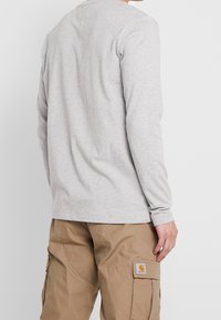 Graues Langarmshirt mit rundem Ausschnitt, leicht strukturiertem Stoff, getragen mit beige Cargo-Hosen, die ein Patch-Logo an der Seite haben.