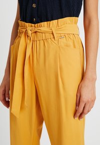 Gula högmidjade byxor med en knuten bälte, med sidofickor och en mjuk textur, i kombination med en ribbad marinblå topp.