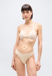 Γυναίκα φοράει μπεζ δαντελωτό τρίγωνο σουτιέν και αντίστοιχα σλιπ με ψηλή μέση, στέκεται μπροστά σε απλό ανοιχτό γκρι φόντο.