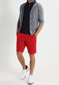 Cardigan gris avec bord noir, polo bleu marine, shorts rouges, et baskets blanches avec accents noirs. Tenue simple et décontractée.