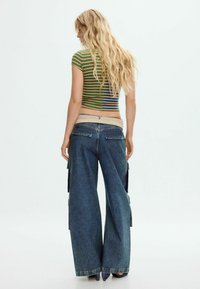 Žalias ir oranžinis juostuotas trumpas viršus, derinamas su plačiomis mėlynomis džinsinio audinio krovinių kelnėmis, kurios turi šonines kišenes ir smėlio spalvos juosmenį.