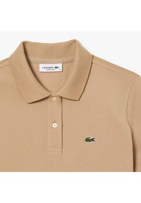 Tan-paitainen polo-paita, joka on valmistettu teksturoituneesta kankaasta, sisältää kolmen napin listan ja vasemmassa rinnassa on brodeerattu vihreä krokotiililogot.