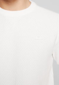 Biały sweter o teksturowanej powierzchni z rombowym wzorem, z okrągłym dekoltem i subtelnym logo po lewej stronie.