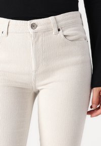 Beige manchestersbyxor med hög midja som har en vertikal ribbad textur, framfickor och en silverfärgad knappstängning i midjan.