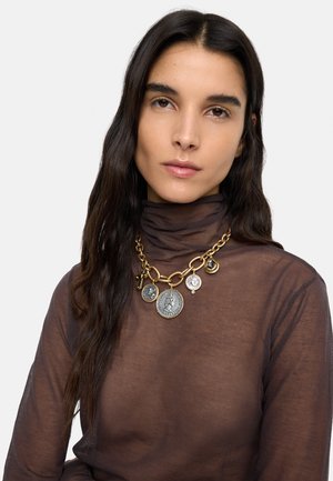 Kobieta o długich ciemnych włosach nosi przezroczysty brązowy golf oraz masywny złoty łańcuch z pięcioma dużymi zawieszkami przypominającymi monety.