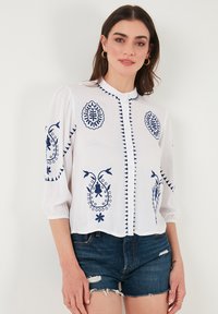 Chemisier blanc avec broderie bleue comportant des motifs floraux et géométriques, manches bouffantes courtes et un design à boutons sur le devant.