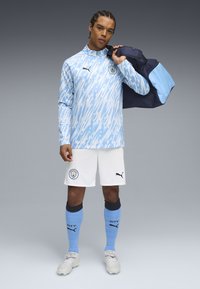 Blaues und weißes langärmeliges Sportoberteil mit abstraktem Druck, weiße Shorts, blaue Socken und graue Sportschuhe. Zubehör umfasst eine blaue Tasche.