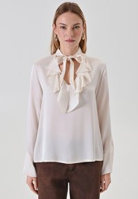 Blusa in seta color crema con scollatura a V arricciata e dettaglio con fiocco. Presenta maniche lunghe con una superficie liscia e una vestibilità comoda.