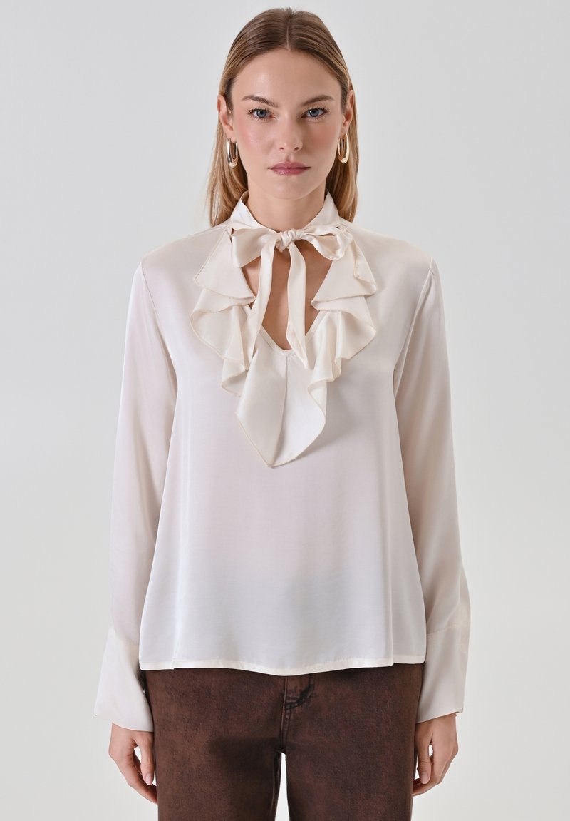 Blusa in seta color crema con scollatura a V arricciata e dettaglio con fiocco. Presenta maniche lunghe con una superficie liscia e una vestibilità comoda.