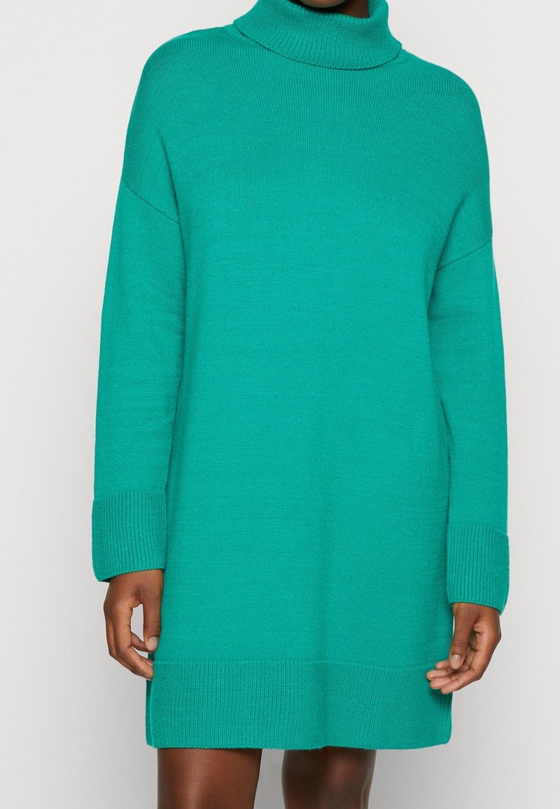 Femme portant une robe-pull tricotée vert sarcelle à manches longues avec un col roulé et des poignets et un ourlet côtelés.