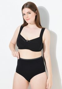 Vrouw met lang bruin haar die een zwarte ondersteunende beha en hoge zwarte onderbroek draagt, staand tegen een witte muur.