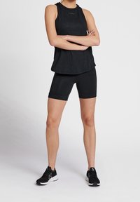 Person som står med armarna i kors, iklädd en svart ärmlös topp, svarta cykelbyxor och svarta träningsskor mot en enfärgad bakgrund.