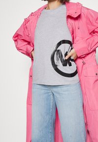 Grijze t-shirt met een groot zwart logo, gelaagd onder een felle roze waterdichte jas. Blauwe wijde jeans maken de outfit compleet.