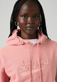 Sudadera rosa con capucha ajustable, que presenta un logotipo en relieve "Levi's 73" en la parte frontal. Tejido suave con una textura lisa.
