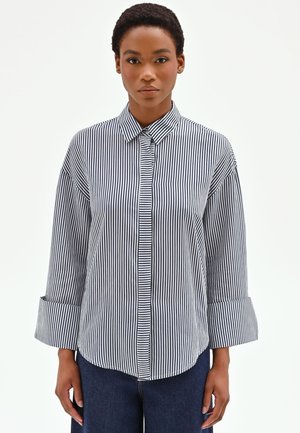 Donna con capelli corti che indossa una camicia a righe verticali bianche e nere con bottoni, polsini larghi e pantaloni blu scuro su sfondo bianco.