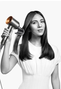 Föhn met een metalen behuizing en koperen accenten, vastgehouden door een persoon die lang, stijl haar stylet. Een witte textuurjurk versterkt de aandacht op het product.