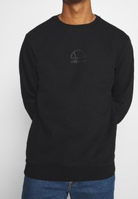 Sudadera negra de cuello redondo con un sutil logo en relieve en el pecho, fabricada en un tejido suave con dobladillo y puños de canalé, combinada con jeans azules.