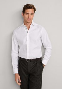 Weißes Hemd mit Knopfleiste, spitzem Kragen, langen Ärmeln und glatter Textur, kombiniert mit dunkelgrauer Hose und braunem Gürtel.