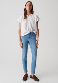 Haut en coton blanc à manches courtes avec un col froncé, assorti à un jean skinny bleu clair. Des chaussures brunes à enfiler complètent la tenue.