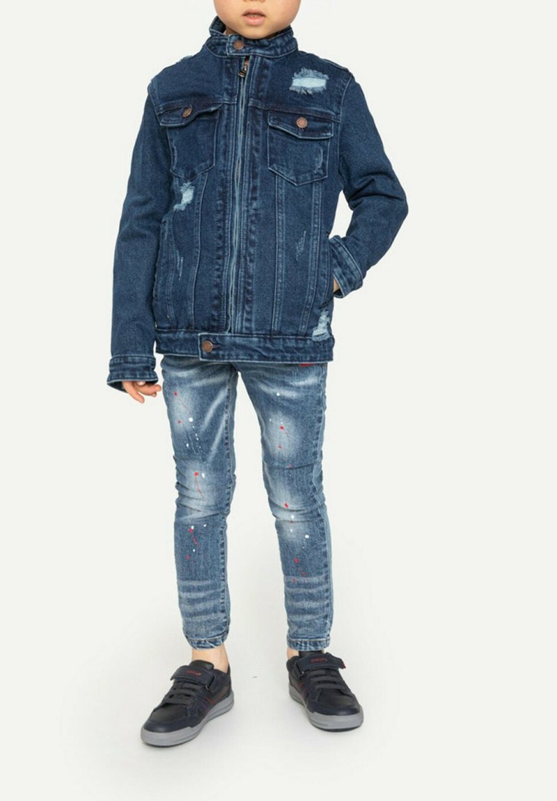 Veste en denim bleu foncé avec deux poches poitrine, détails usés et col haut. Associée à un jean bleu clair avec éclaboussures de peinture et des baskets noires.