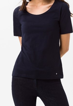 Frau trägt ein schlichtes schwarzfarbenes T-Shirt mit U-Ausschnitt und kurzen Ärmeln sowie schwarze Hosen, die Hände hinter dem Rücken verschränkt.