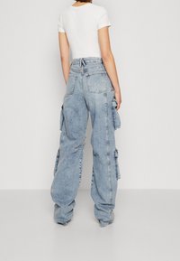 Osoba v světle modrých džínech typu cargo s širokými nohavicemi a velkými bočními kapsami, oblečená v přiléhavém bílém tričku s krátkým rukávem, stojící zády k nám.