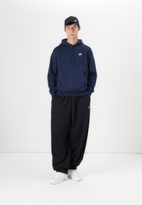 Sweatshirt azul-marinho com um bolso frontal e logótipo branco, combinado com calças de fato pretas largas. Usado com ténis brancos e um boné preto.