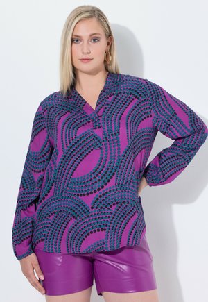 Vibrant purple blouse with black and teal dot patterns, featuring a notched collar and long sleeves, paired with shiny purple shorts.