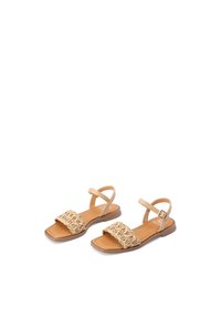 Sandali piatti beige con design a fascetta intrecciata, materiale sintetico e fibbia alla caviglia. Punta aperta con una suola piatta liscia.