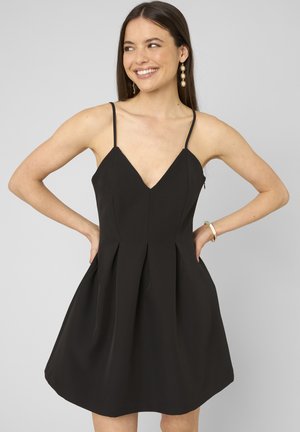 Femme souriante, les mains sur les hanches, portant une robe noire sans manches en V et des boucles d'oreilles dorées en forme de soleil sur fond gris.