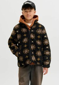 Crna fleeced jakna s šarenim geometrijskim uzorkom po cijeloj površini, s prednjim zatvaračem i bočnim džepovima. Nosi se preko smešnog dukserice.