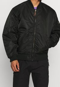 Black bomber jacket with a smooth fabric, ribbed cuffs, elastic waistband, zip closure, and side pockets. Features a gold zipper and contrast lining.
