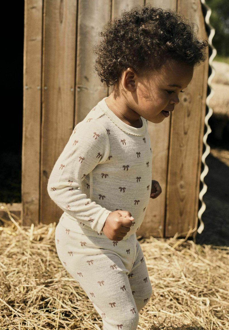 Un jeune enfant aux cheveux bouclés portant un pyjama crème avec de petits nœuds bruns, marchant à l'extérieur près d'une structure en bois sur un sol recouvert de paille.