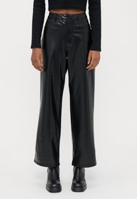 Calças pretas de cintura alta e perna larga feitas de material texturizado brilhante com padrão de diamante. Combinadas com botas de plataforma pretas até ao tornozelo.