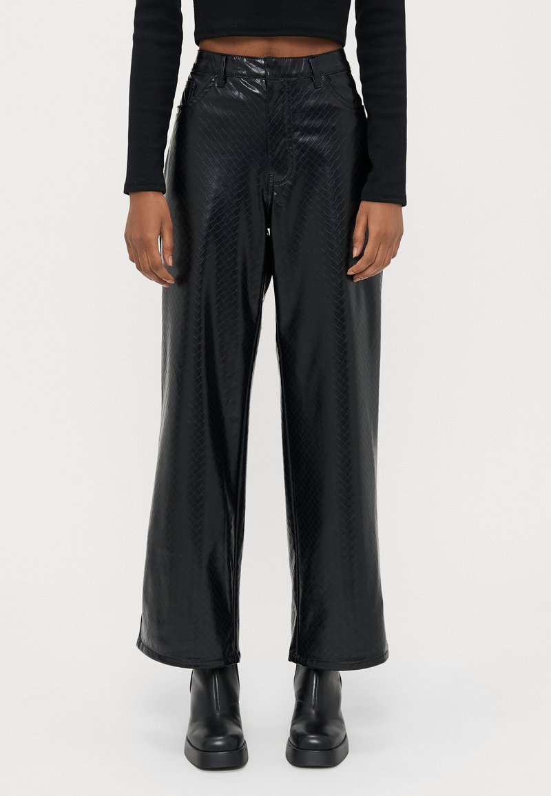 Calças pretas de cintura alta e perna larga feitas de material texturizado brilhante com padrão de diamante. Combinadas com botas de plataforma pretas até ao tornozelo.