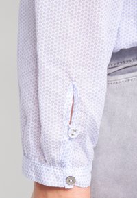 Manche de chemise bleu clair à motifs avec petits cœurs, dotée d'une fermeture par bouton et d'une fente pour la ventilation au niveau du poignet.