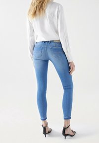 Jeans ajustados de mezclilla en azul claro con una textura suave, que presentan un diseño clásico de cinco bolsillos y sutiles detalles de costura. Combinados con una camisa blanca.