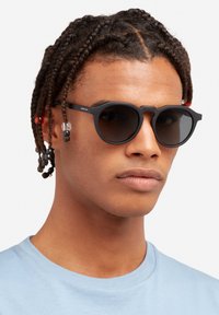 Okrúhle slnečné okuliare s čiernym plastovým rámom, tmavými sklami a šesťuholníkovým tvarom; doplnené farebnými korálkami vo vlasoch. Modelka má na sebe svetlomodrú košeľu.