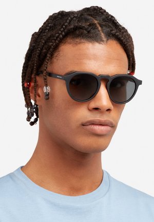 Ringikujulised päikeseprillid musta plastikraamiga, tumedate läätsedega ja kuusnurkse kujuga; juustes on värvilised helmed. Modell kannab helesinist särki.