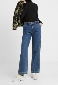 Jean en denim bleu taille haute à coupe large, associé à un col roulé noir côtelé et une veste camouflage. Des baskets blanches complètent la tenue.