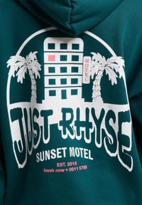 Teal színű kapucnis pulóver, amelyen grafikai design található, fehér felirattal: "JUST RHYSE" és "SUNSET MOTEL". Továbbá pálmafák és egy épület, piros kiemelésekkel.