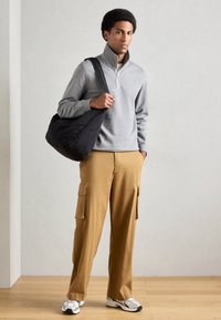 Γκρι φλις μπλούζα με φερμουάρ, καφέ cargo παντελόνια με πλαϊνές τσέπες, μαύρη τσάντα ώμου και λευκά αθλητικά παπούτσια με γκρι και μαύρες λεπτομέρειες.