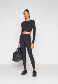 Ensemble de vêtements de sport noirs comprenant un crop top à manches longues et des leggings taille haute, tous deux ornés de bandes avec logo. Une personne tient un sac de sport noir.