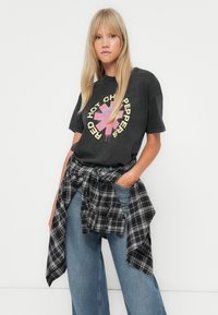 Black graphic t-shirt featuring a circular design with pink and yellow accents, paired with blue jeans and a black and white plaid shirt tied around the waist.