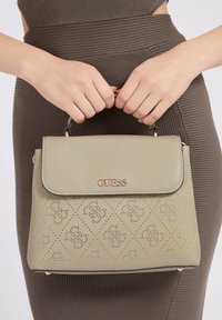 Oliveenvihreä teksturoitu käsilaukku, jossa on kullatut metalliosat, läpällinen suljenta ja kohokuvioitu timanttimalli. Kahvaa pitelee kaksi kättä.